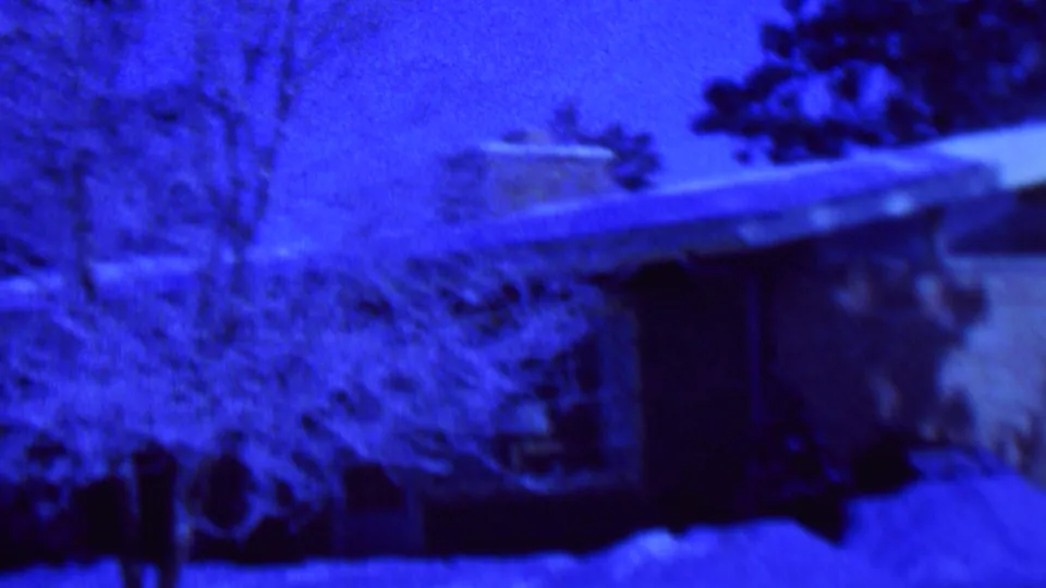 Blue-shifted 1962 St. Paul home-movie frame showing a low house, chimney, bare tree, shrubs, and heavy snow across the foreground.