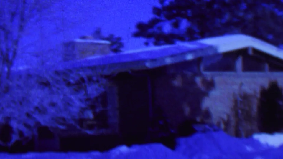 Blue-tinted 1962 8mm frame from St. Paul, Minnesota, showing a snow-covered suburban brick house with a sloped roof, trees, shrubs, and deep snow in the yard.