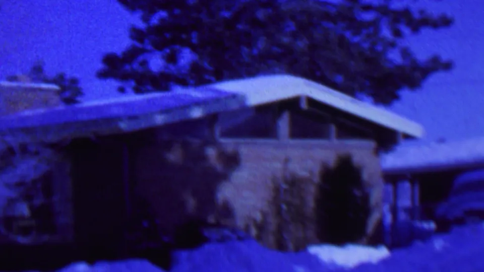 Blue-tinted 1962 St. Paul frame showing a snow-covered mid-century house exterior, shrubs along the wall, deep snow, trees overhead, and a partial vehicle near the garage area.