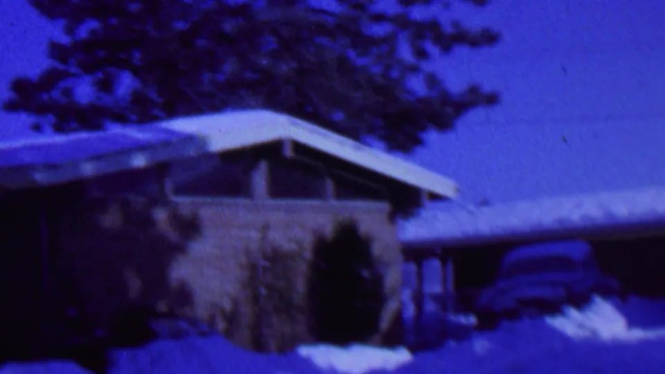 Blue-tinted 1962 St. Paul home-movie frame showing the snowy brick house, attached garage or carport, partially visible older car, shrubs, trees, and heavy snow cover.
