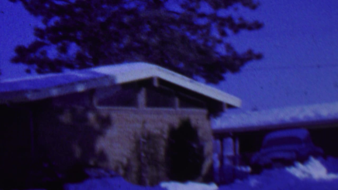 The 1962 St. Paul home-movie frame shows a low modern house with a snow-covered roof, dark entry area, evergreen trees, and a parked car partly obscured by snow.