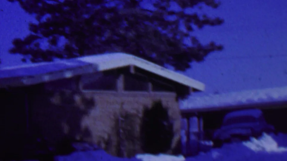 The 1962 St. Paul home-movie frame shows a low modern house with a snow-covered roof, dark entry area, evergreen trees, and a parked car partly obscured by snow.