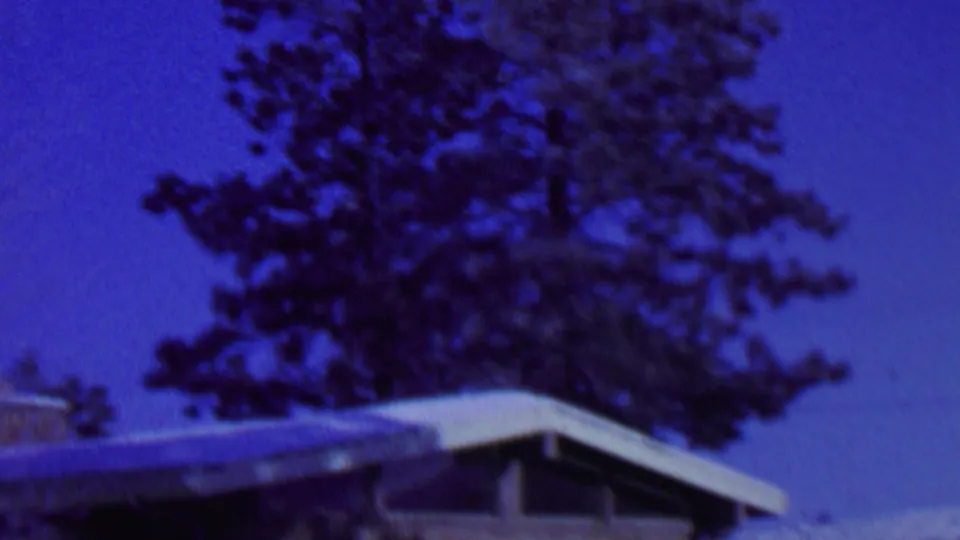 The final frame holds on the snow-covered roofline of the St. Paul residential exterior, with tall evergreen trees behind the house and strong blue 8mm color shift.