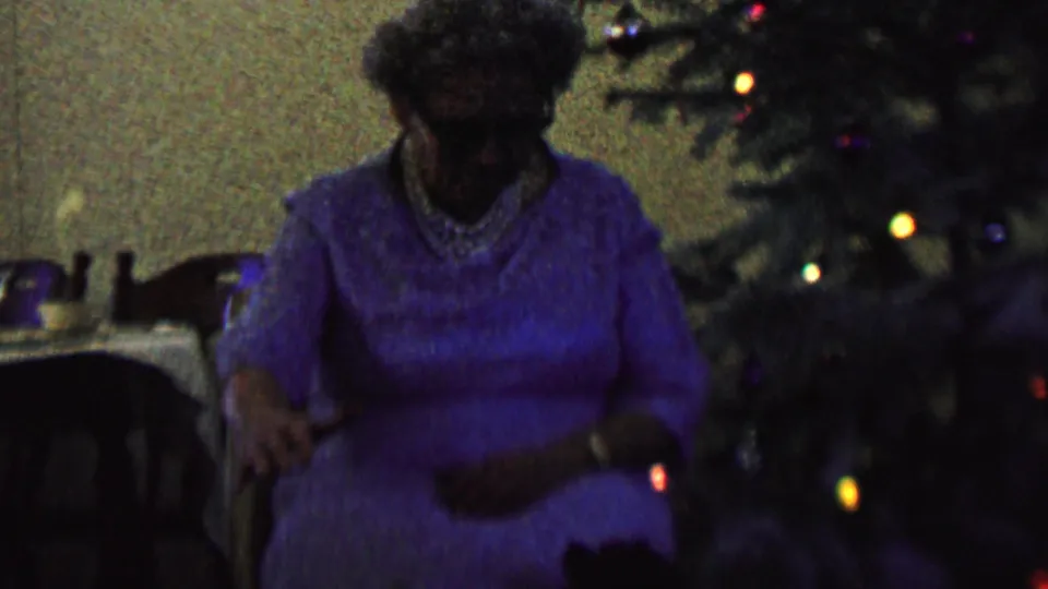 A woman in a purple dress sits in a private interior beside a decorated Christmas tree with small glowing lights, matching the 1962 home-movie holiday metadata.