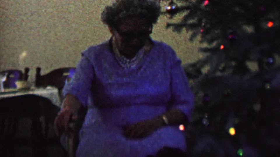 In 1962 St. Paul, Minnesota home-movie footage, an elderly woman in a blue dress sits in a chair beside a decorated Christmas tree with colored lights.