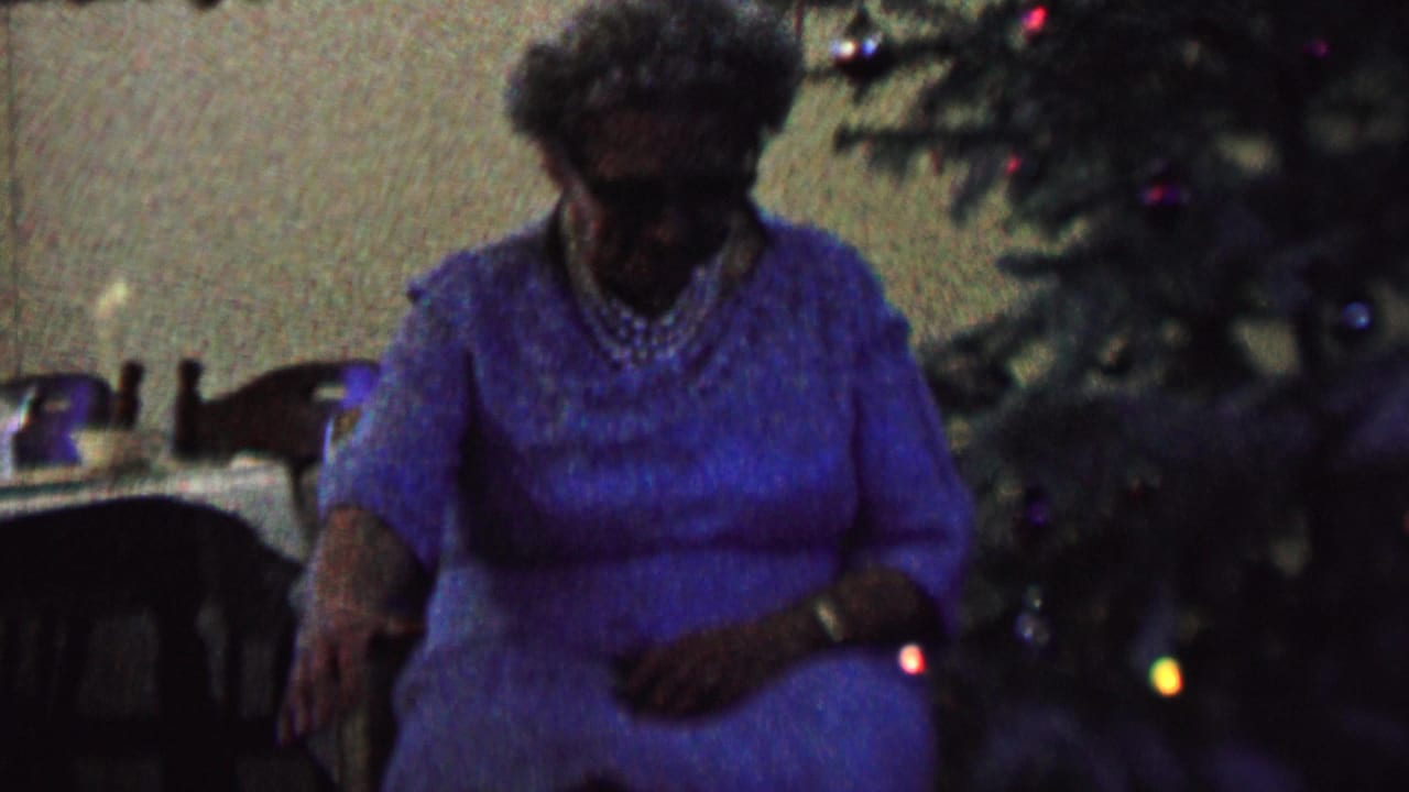 The elderly woman remains seated near the Christmas tree in the St. Paul home interior, with her hands low and her face partly shadowed by the dim exposure.