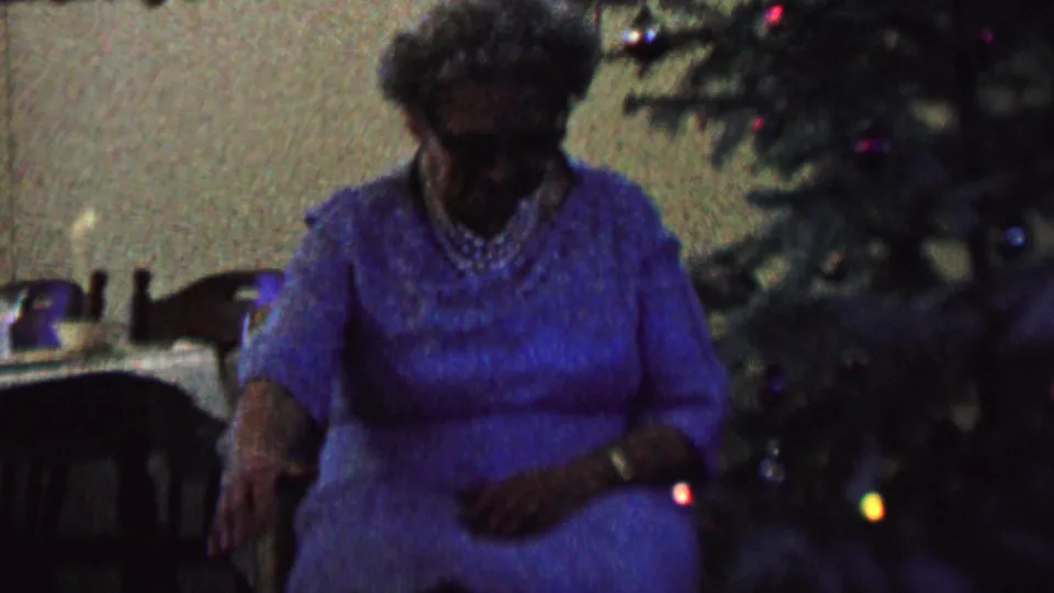 The elderly woman remains seated near the Christmas tree in the St. Paul home interior, with her hands low and her face partly shadowed by the dim exposure.