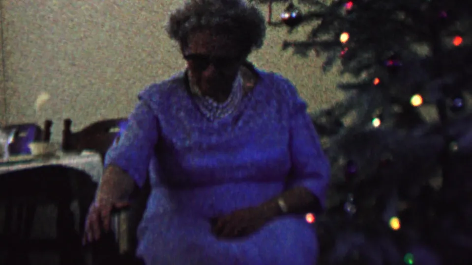 A 1962 home-movie frame shows the elderly woman in blue leaning slightly forward near the chair arm while the Christmas tree and holiday lights fill the right side of the frame.