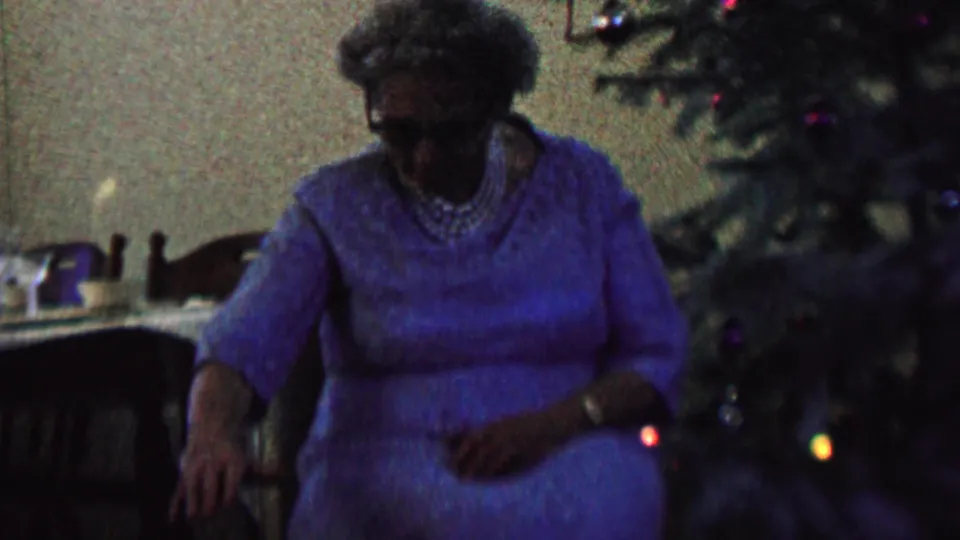 The final frame keeps the woman seated beside the decorated tree in the private St. Paul interior, with colored lights visible against the dark, grainy 8mm image.
