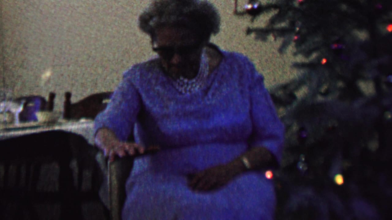 In a 1962 home-movie frame from the supplied St. Paul, Minnesota metadata, an elderly woman wearing a blue dress and pearls sits near a decorated Christmas tree while a dark dog is partly visible in front of her.