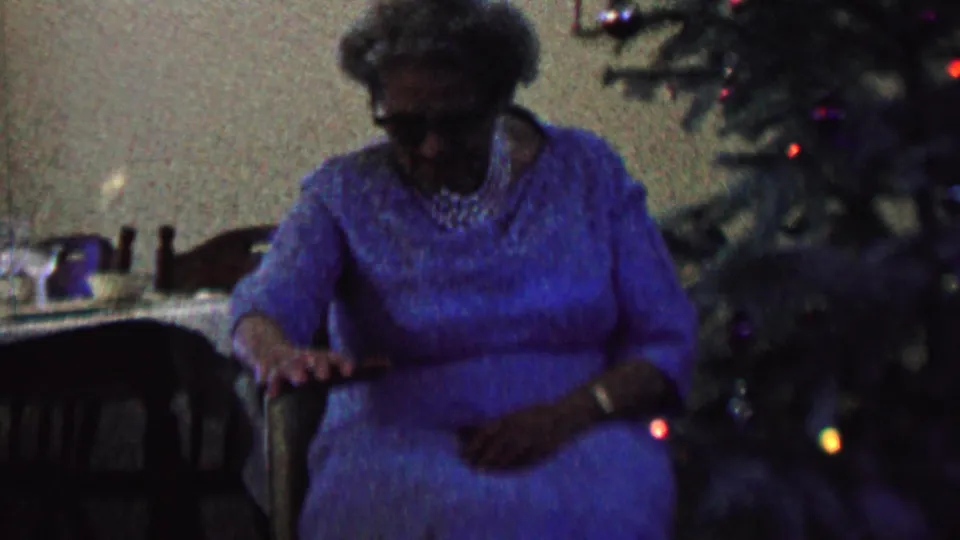 In a 1962 home-movie frame from the supplied St. Paul, Minnesota metadata, an elderly woman wearing a blue dress and pearls sits near a decorated Christmas tree while a dark dog is partly visible in front of her.
