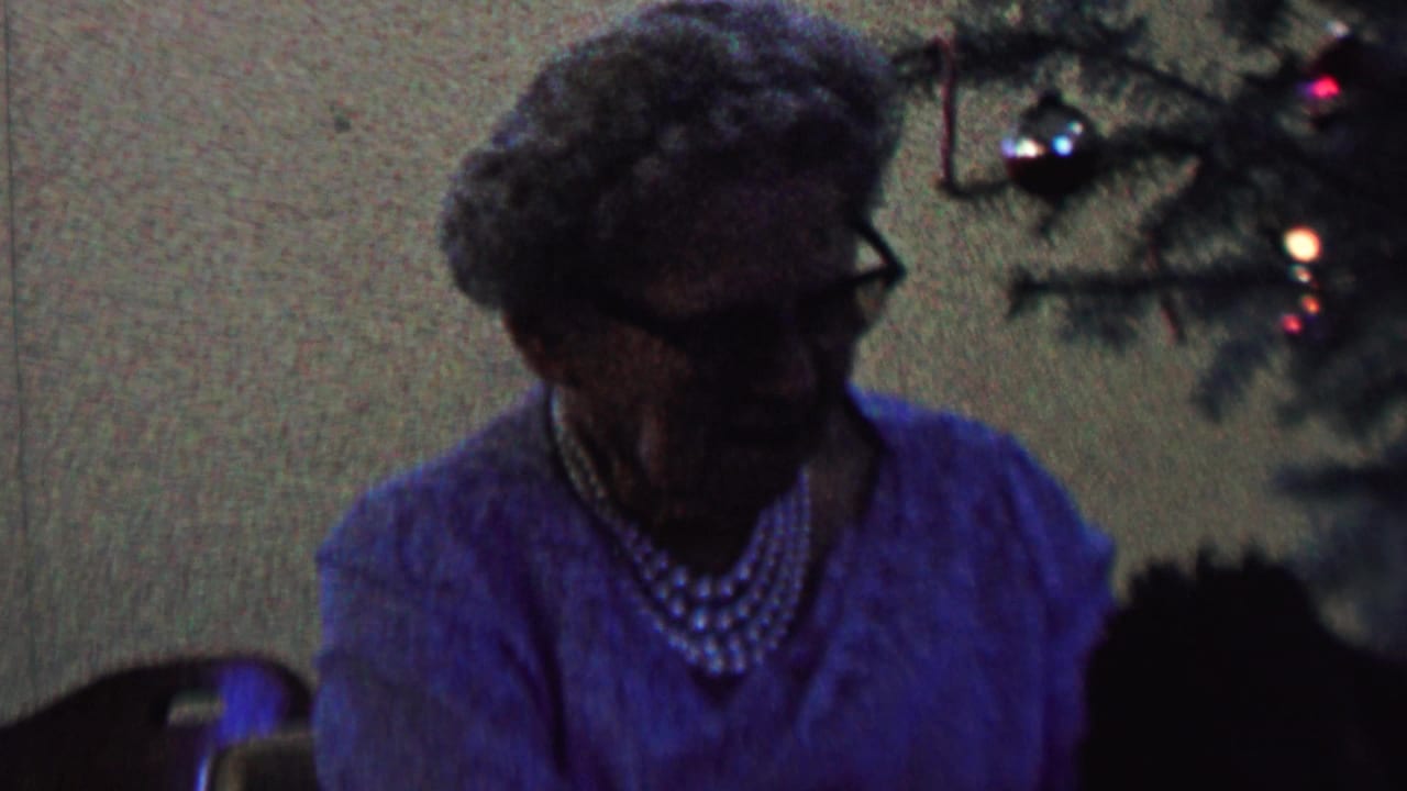 Elderly woman in glasses and pearl necklace beside a Christmas tree.