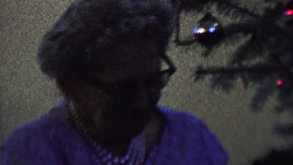 The elderly woman turns in profile near Christmas tree branches and reflective ornaments, with her glasses and pearl necklace visible in the dim 8mm frame.