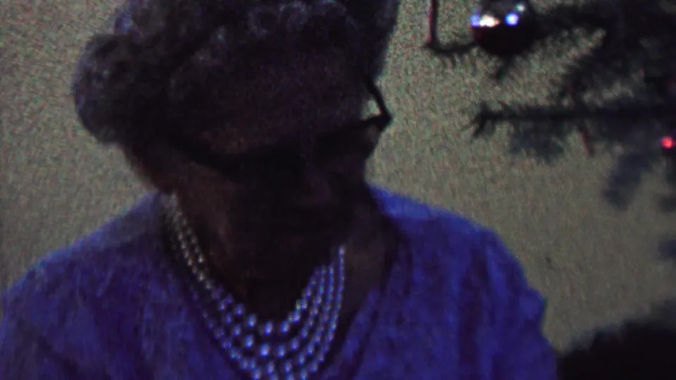 Close, shadowed 8mm view of the woman in glasses and pearls looking toward a Christmas tree branch with a small colored light in the background.