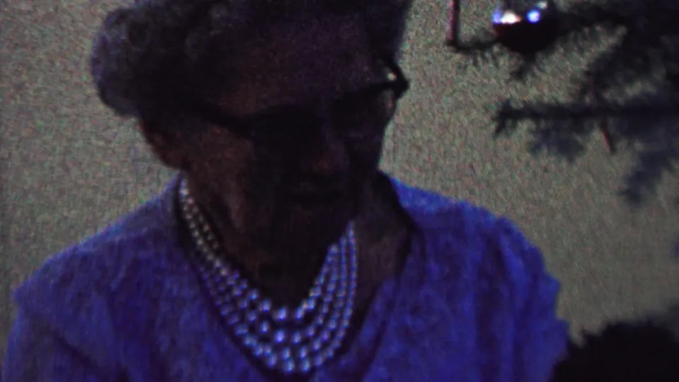 The woman faces slightly toward camera in a blue sweater and layered pearl necklace, with Christmas tree branches and ornaments behind her in the St. Paul home interior.
