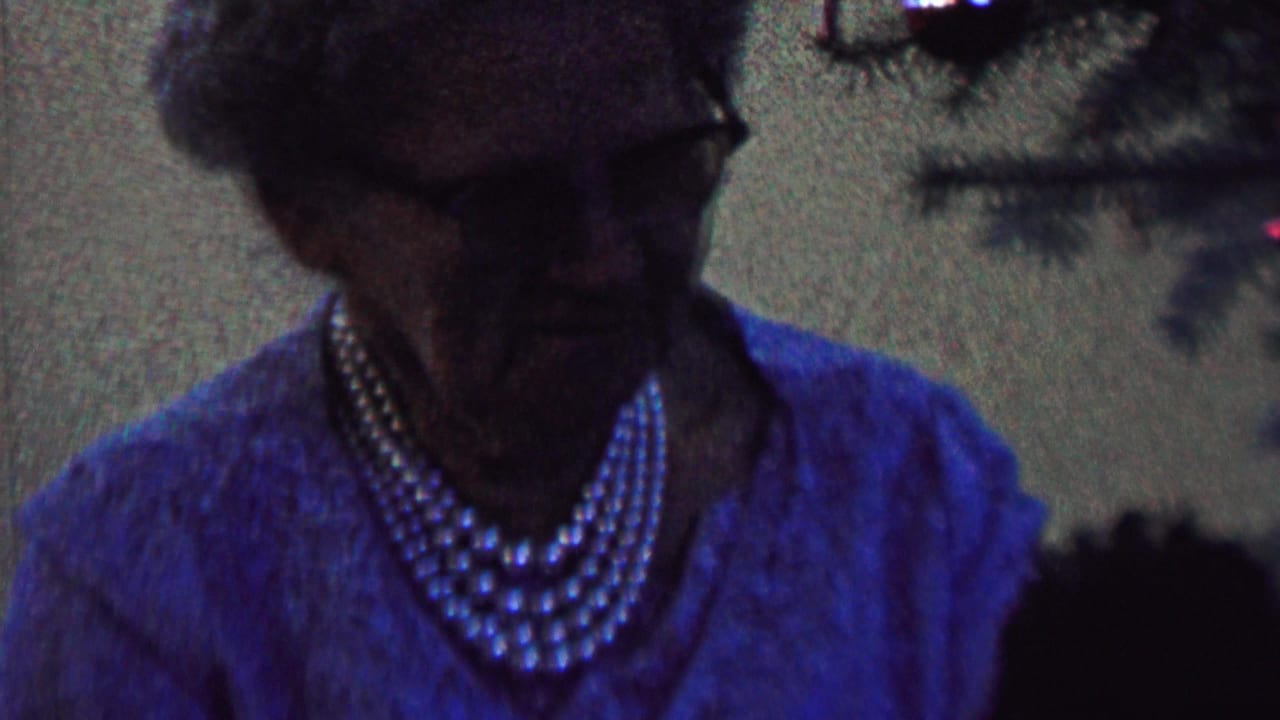 Elderly woman with gray hair, glasses, blue clothing, and layered pearls seated near a decorated Christmas tree in archival home-movie footage from the supplied St. Paul, Minnesota 1962 metadata.