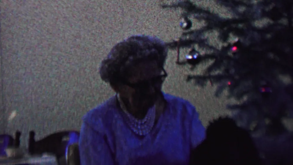 A wider interior frame shows the elderly woman seated near the Christmas tree, with ornaments, chair details, and household furnishings visible in the low-light 8mm image.