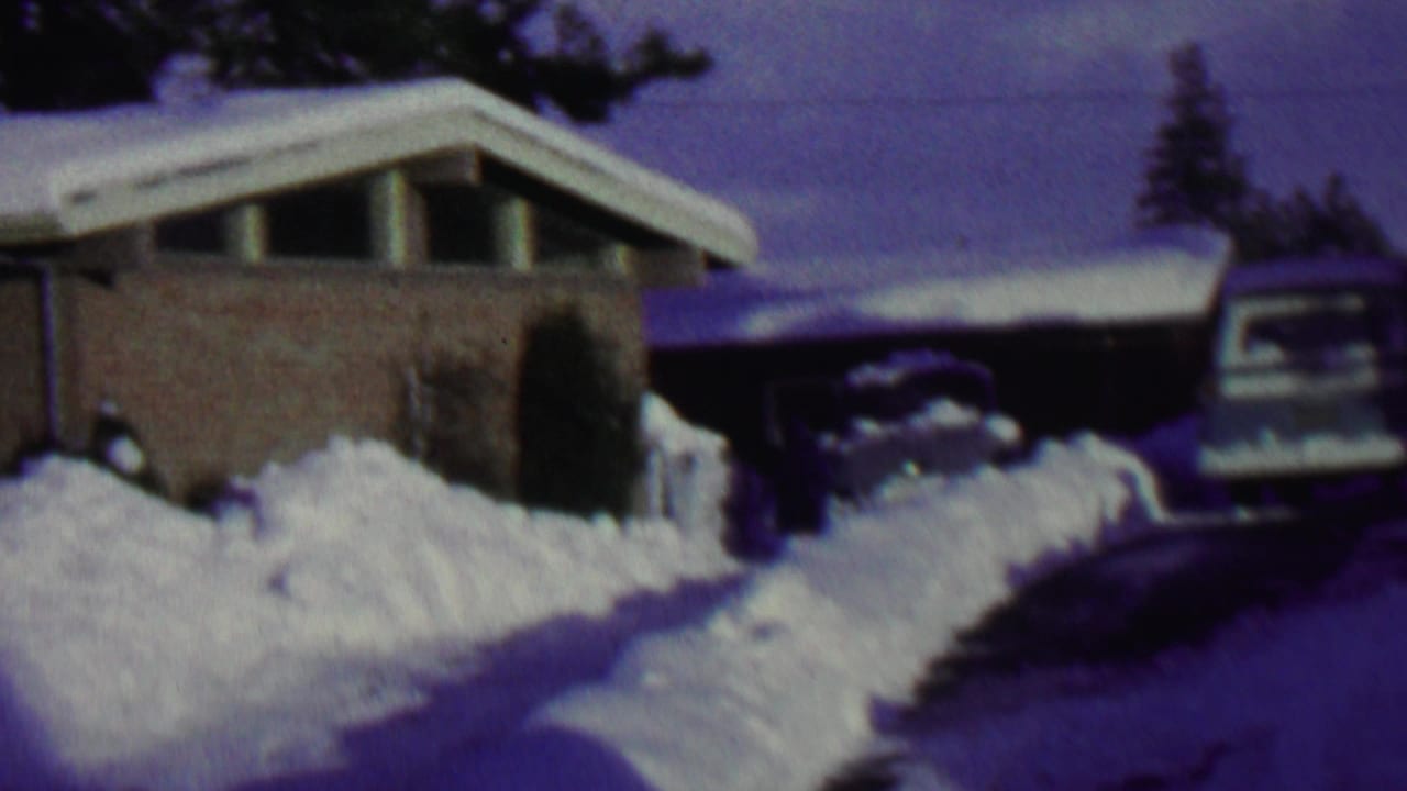 1962 St. Paul home-movie frame showing a low brick suburban house, deep snowbanks, a plowed driveway, and a car partly surrounded by snow.