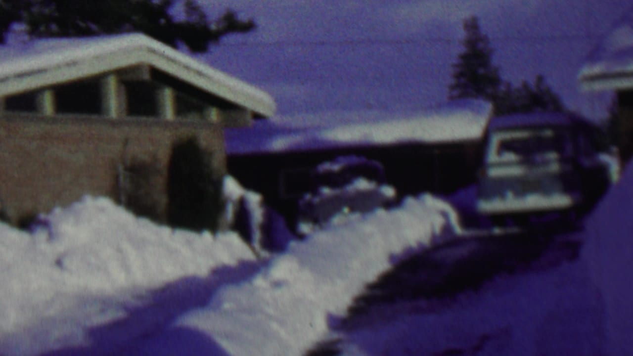 1962 St. Paul frame of a snow-laden residential driveway, with a brick house on the left, a car near the garage, and heavy snow piled along the drive.