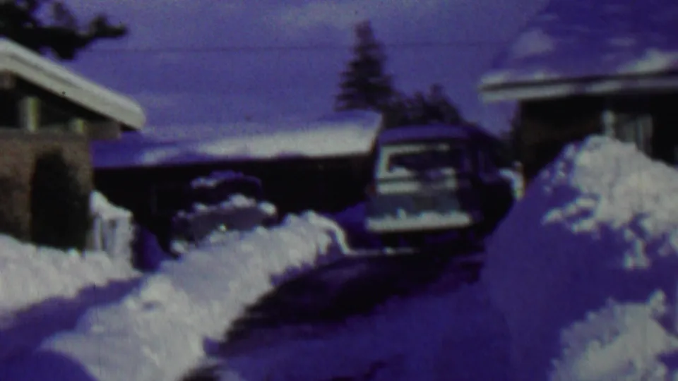 1962 St. Paul home-movie view across high snowbanks toward a residential driveway, a parked car, and neighboring house roofs under a blue winter cast.