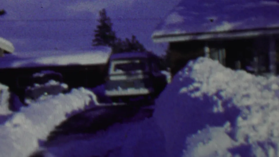 1962 St. Paul frame showing a large foreground snowbank, a driveway curving toward a parked car, and snow-covered suburban houses in soft 8mm focus.