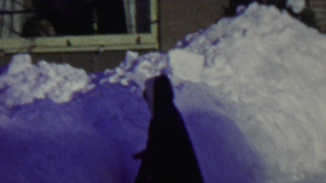 In 1962 St. Paul home-movie footage, a dark hooded figure stands near deep piled snow outside a residential window.