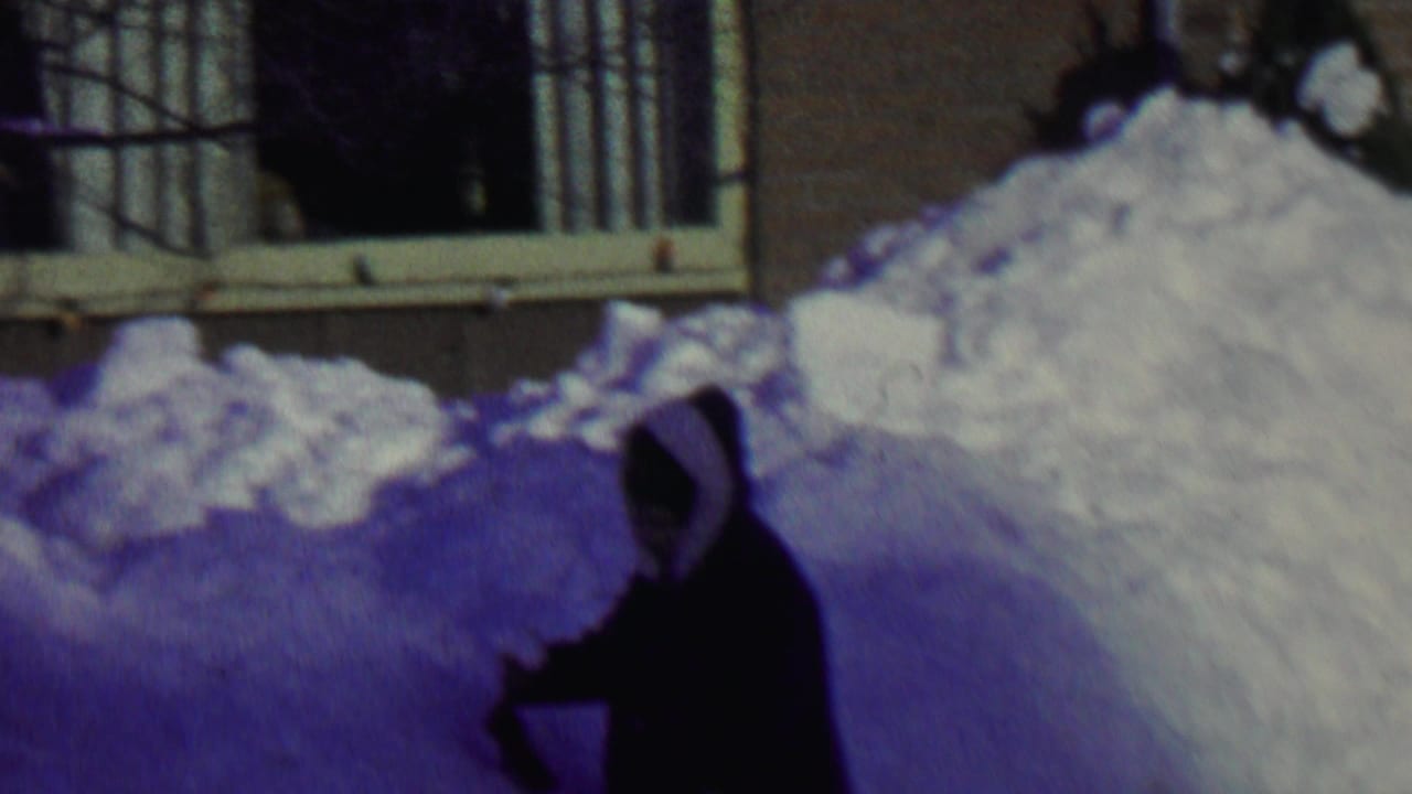 A person in a dark hooded winter coat appears to shovel deep snow beside a house in the 1962 St. Paul clip.