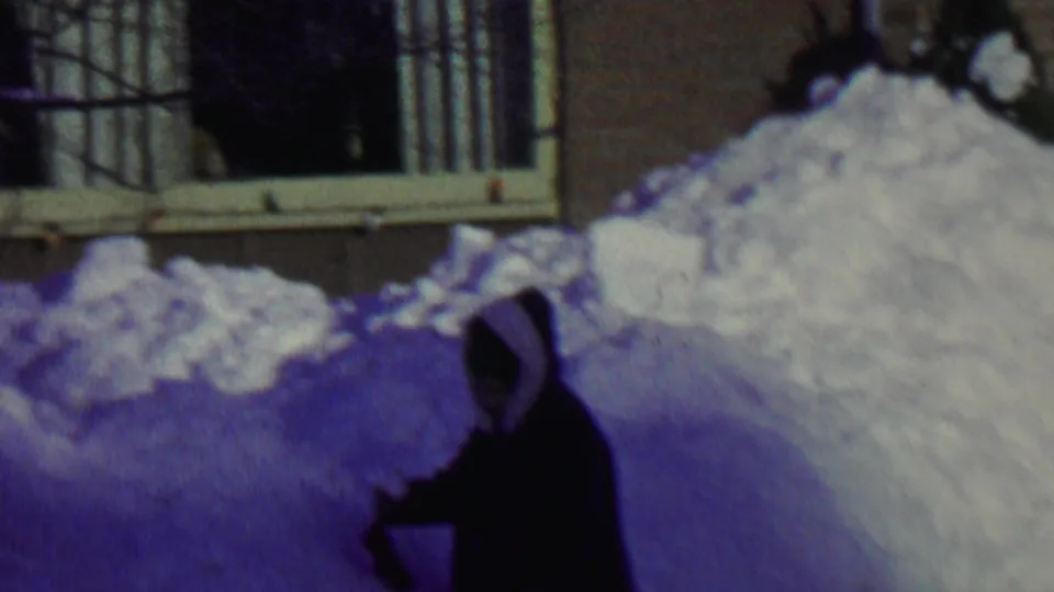 A person in a dark hooded winter coat appears to shovel deep snow beside a house in the 1962 St. Paul clip.