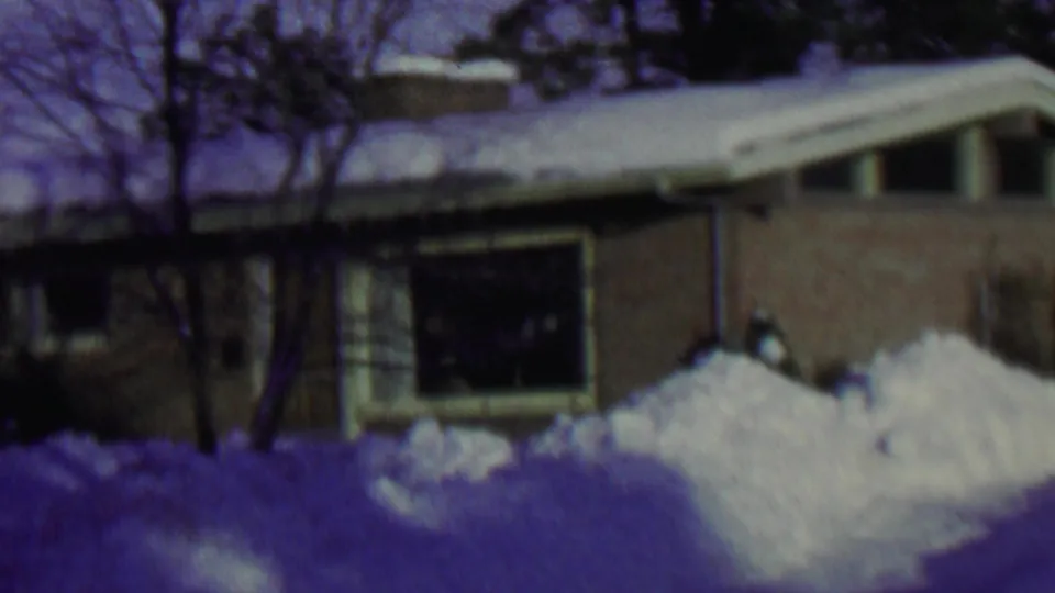 1962 St. Paul home-movie frame of a low brick residential house with heavy roof snow, bare trees, and deep snow piled near the front window.