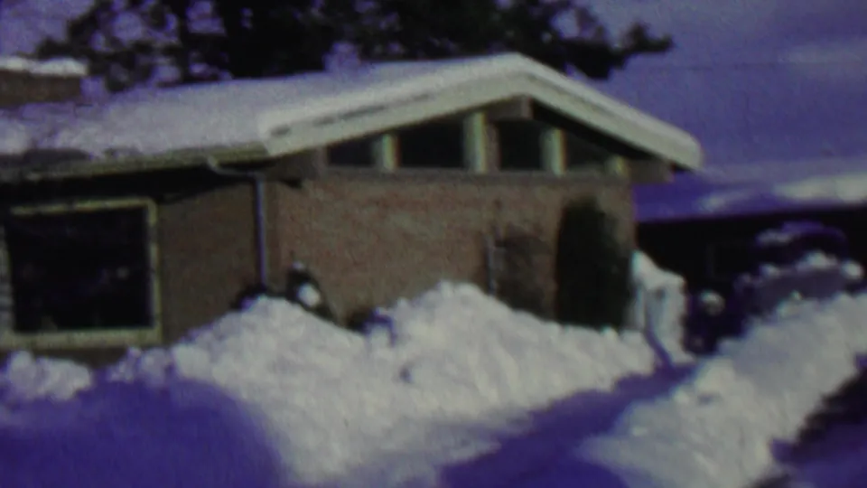 1962 St. Paul winter frame showing a brick home with roof snow, a dark front window, piled snow, and a partially cleared walkway or driveway edge.