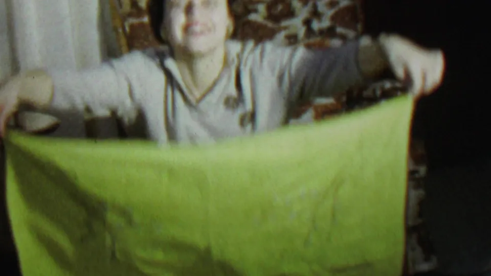 A woman in a light-colored sweater holds a bright green sheet in a private living room, with patterned seating and curtains behind her.