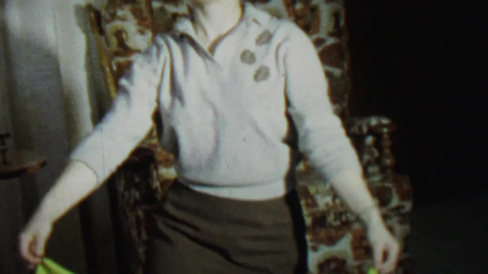The woman stands in front of a patterned chair after lowering or dropping the green sheet, continuing the playful home-movie trick.