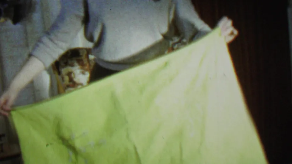 The woman bends or leans while holding the green sheet near a patterned chair in the same private living-room setting.