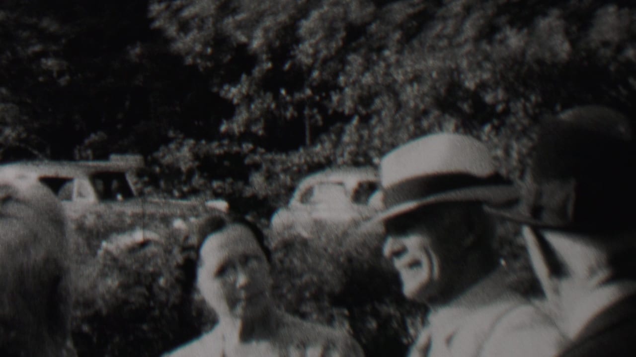 A man in a brimmed hat faces another adult while a woman stands nearby outdoors.