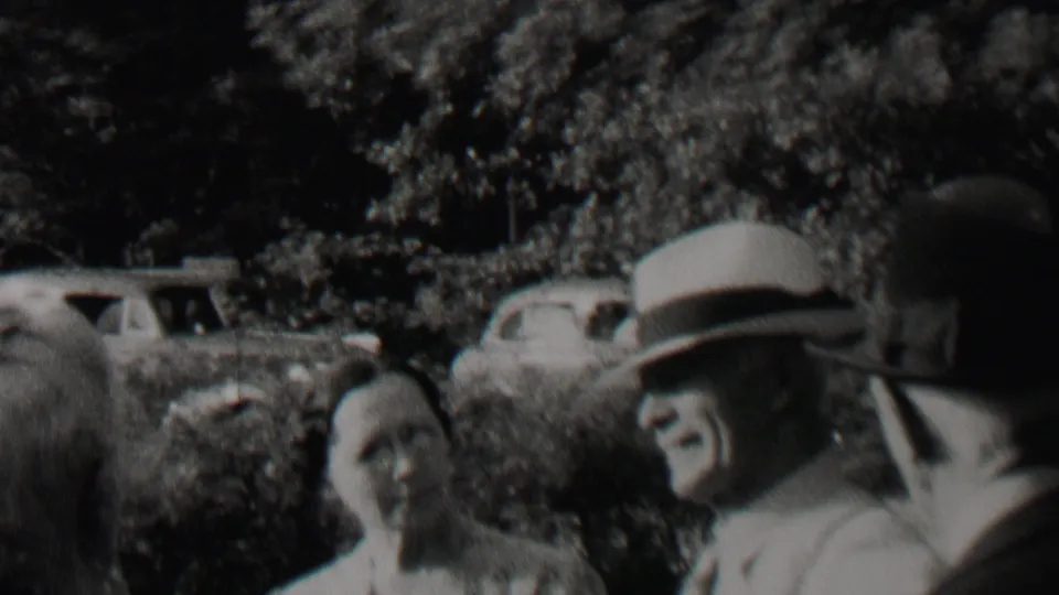 A close 1937 home-movie view shows a man in a brimmed hat and suit turned toward another adult, with a woman and parked automobiles visible in the background.