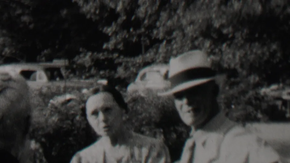 A woman in light clothing and a man in a brimmed hat face the home-movie camera outdoors, with foliage and parked automobiles behind them in the supplied 1937 Trenton context.