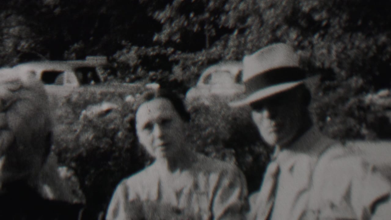 Black-and-white 8mm frame from the supplied 1937 Trenton, New Jersey clip showing older adults outdoors near bushes, with period cars visible beyond them.