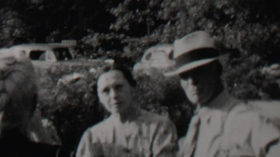 Black-and-white 8mm frame from the supplied 1937 Trenton, New Jersey clip showing older adults outdoors near bushes, with period cars visible beyond them.