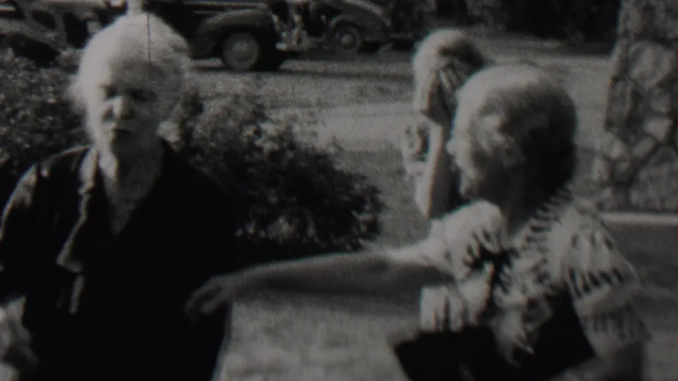 Black-and-white 8mm frame showing an elderly woman in a dark dress facing the camera as another person at right raises an arm in the outdoor residential setting.