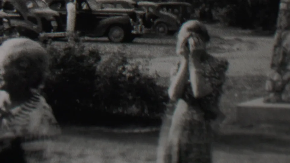 Black-and-white 8mm frame from the supplied Trenton home movie showing people in an outdoor yard or sidewalk area, including one person covering their face near parked 1930s automobiles.