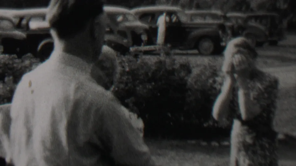 Black-and-white 8mm frame showing a man from behind in the foreground, with other people partly visible near bushes and parked cars in the supplied 1937 Trenton clip.