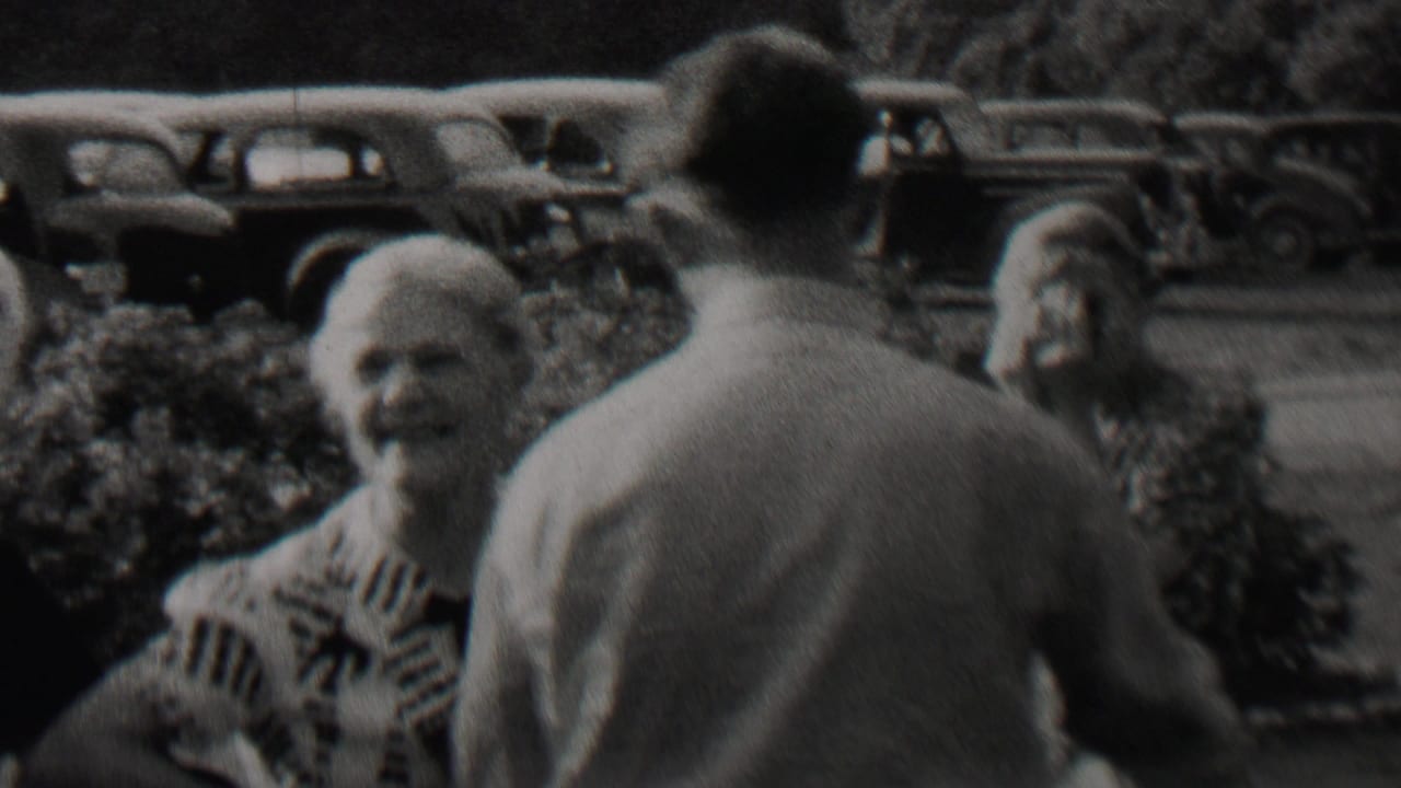 1937 Trenton, New Jersey home-movie frame showing older adults talking outdoors, with hedges and parked 1930s automobiles in the background.
