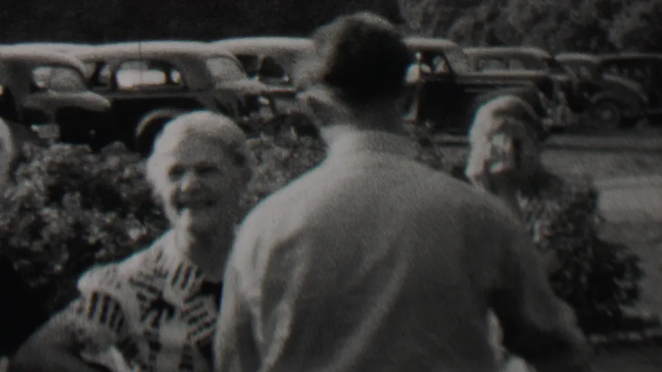 1937 Trenton, New Jersey home-movie frame showing older adults talking outdoors, with hedges and parked 1930s automobiles in the background.