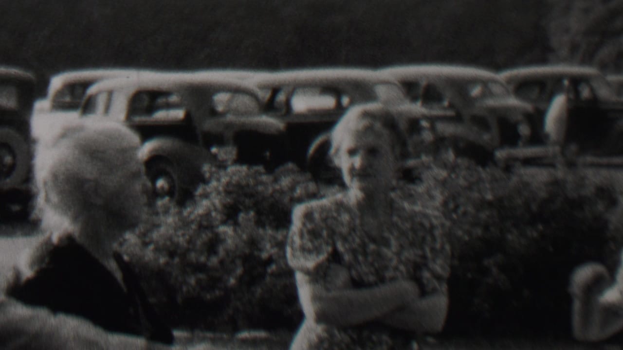 Two older women and a man stand near hedges with parked cars behind them.