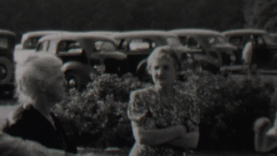 1937 Trenton, New Jersey frame with older women in dresses and a suited man at the edge of the shot, set against hedges and parked automobiles.