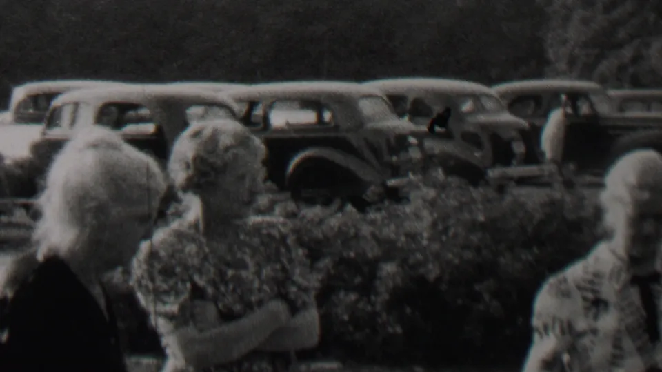 1937 Trenton, New Jersey home-movie frame showing older adults in conversation, with a woman in a patterned dress and a row of cars behind the hedge.