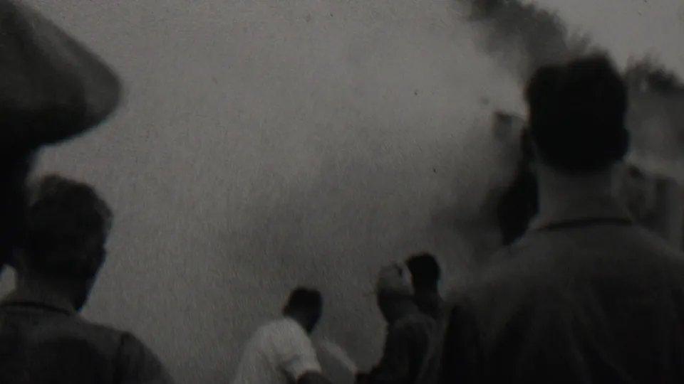 From within the gathered crowd, the camera shows men standing near thick smoke or fumes, with the scene softened by grain and handheld 8mm exposure.