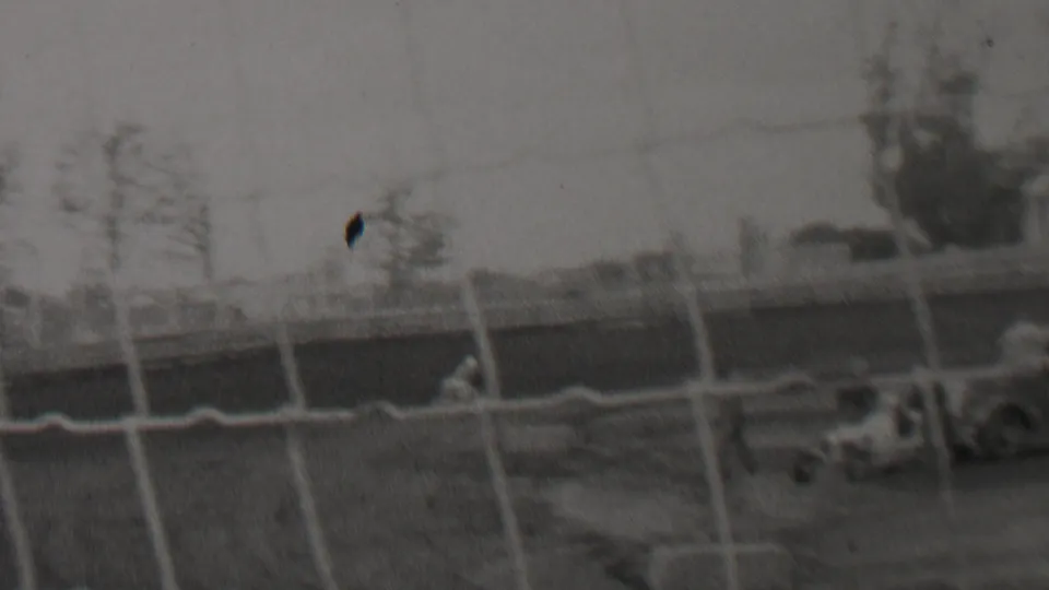 A vintage open-wheel race car moves along the dirt track in the supplied 1937 Trenton, New Jersey home-movie footage, seen through a chain-link fence with distant figures and poles behind it.