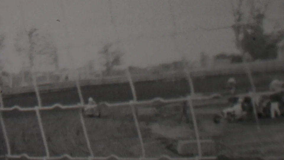 Several low race cars and small trackside figures appear beyond a chain-link fence in the 1937 Trenton racing footage, with trees and a hazy background across the track.