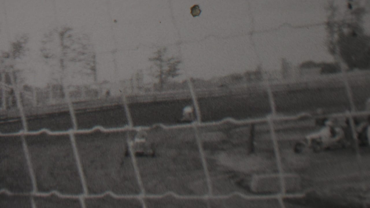 A small racing car appears behind protective netting on an outdoor dirt track in archival 1937 United States home-movie footage.